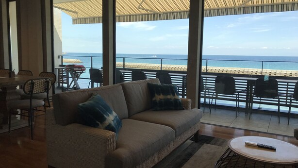 A living room with a sofa, chairs, and a table. There is a large window with a view of the sea and a beach. - Villa Ocean Kamaguchi (Awaji)