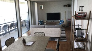 A modern living room with a sofa, a dining table, and a TV. There is a balcony with chairs and a railing. - Villa Ocean Kamaguchi (Awaji)