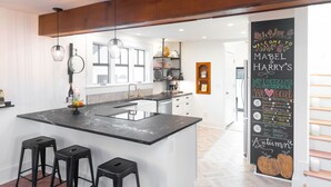 A modern kitchen with a large island, bar stools, pendant lights, and a chalkboard wall with menu items. - Mabel and Harry's, a modern country guest house (Kokomo)
