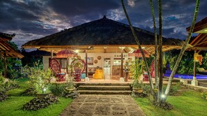 A traditional thatched-roof building with a veranda, surrounded by lush greenery and a pool.