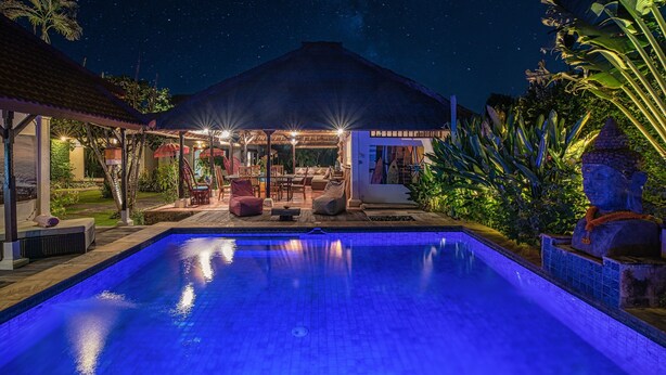 A swimming pool with a night sky view, a covered seating area, and tropical plants.