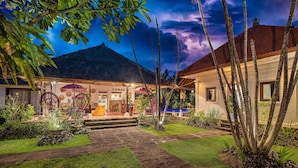 A well-lit building with a thatched roof, surrounded by lush greenery and palm trees.