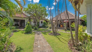 A tropical garden with a stone pathway leading to a house with a thatched roof.