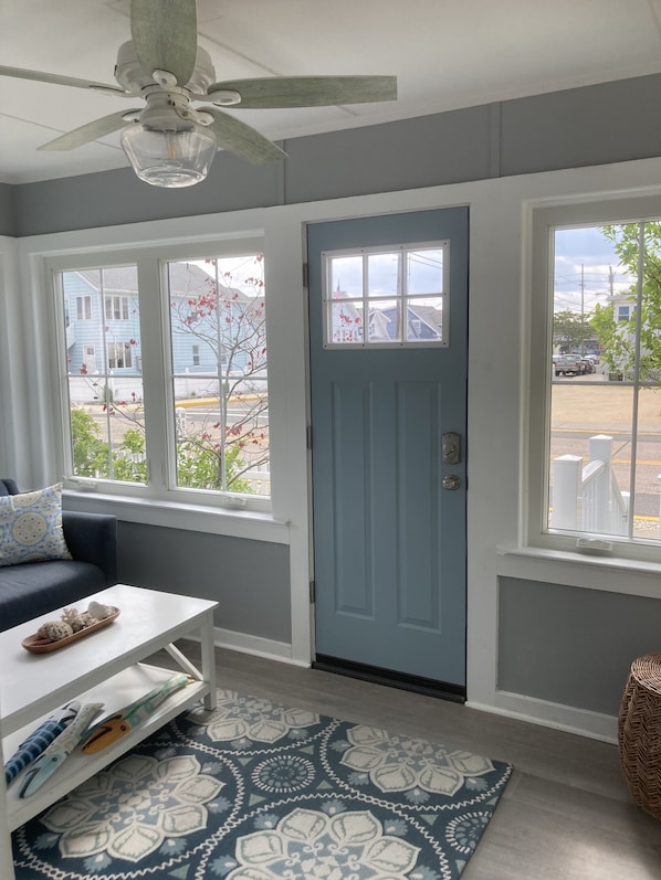 A room with a blue door, a ceiling fan, a window with a view of houses, a patterned rug, and a white table with items on it.