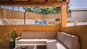 Un espace extérieur aménagé, abrité, avec un canapé et une table, un mur en pierre et une vue sur une piscine bordée de chaises bleues.