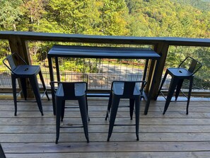 A wooden deck with a metal table and four metal chairs, overlooking a forested area.