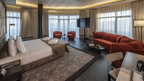 Ett modernt hotellrum med en stor säng, en röd soffa, två fåtöljer, en platt-TV och stora fönster med gardiner.