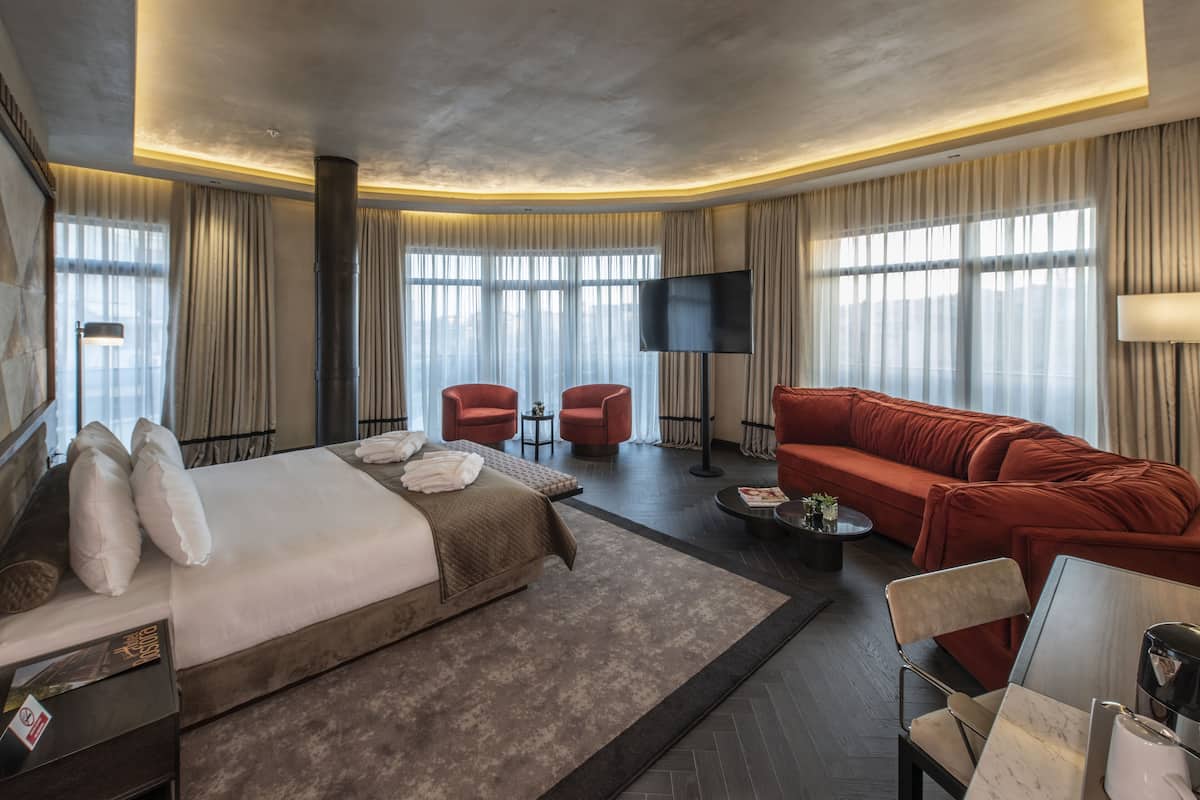 Una habitación de hotel moderna con una cama grande, un sofá rojo, dos sillones, un televisor de pantalla plana y ventanas amplias con cortinas.