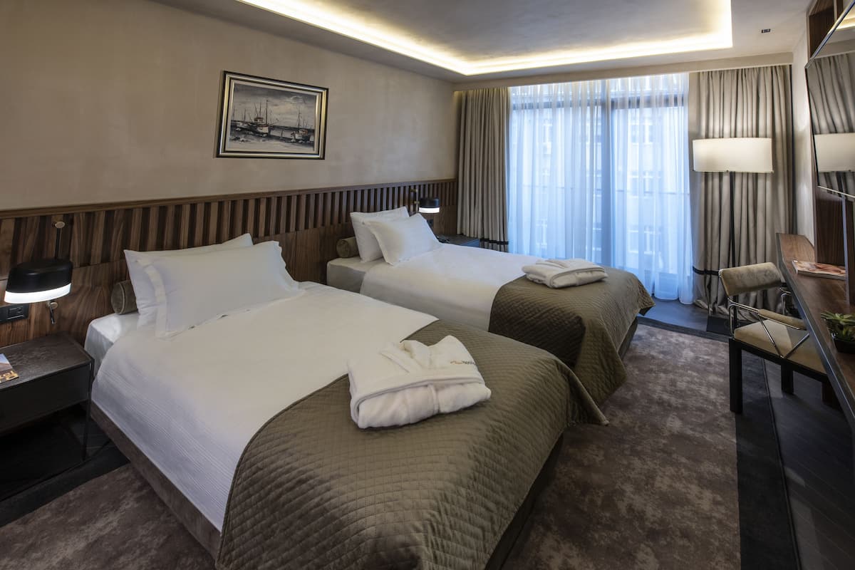 Habitación de hotel con dos camas, un escritorio, una silla, una lámpara y un ventanal con cortinas.