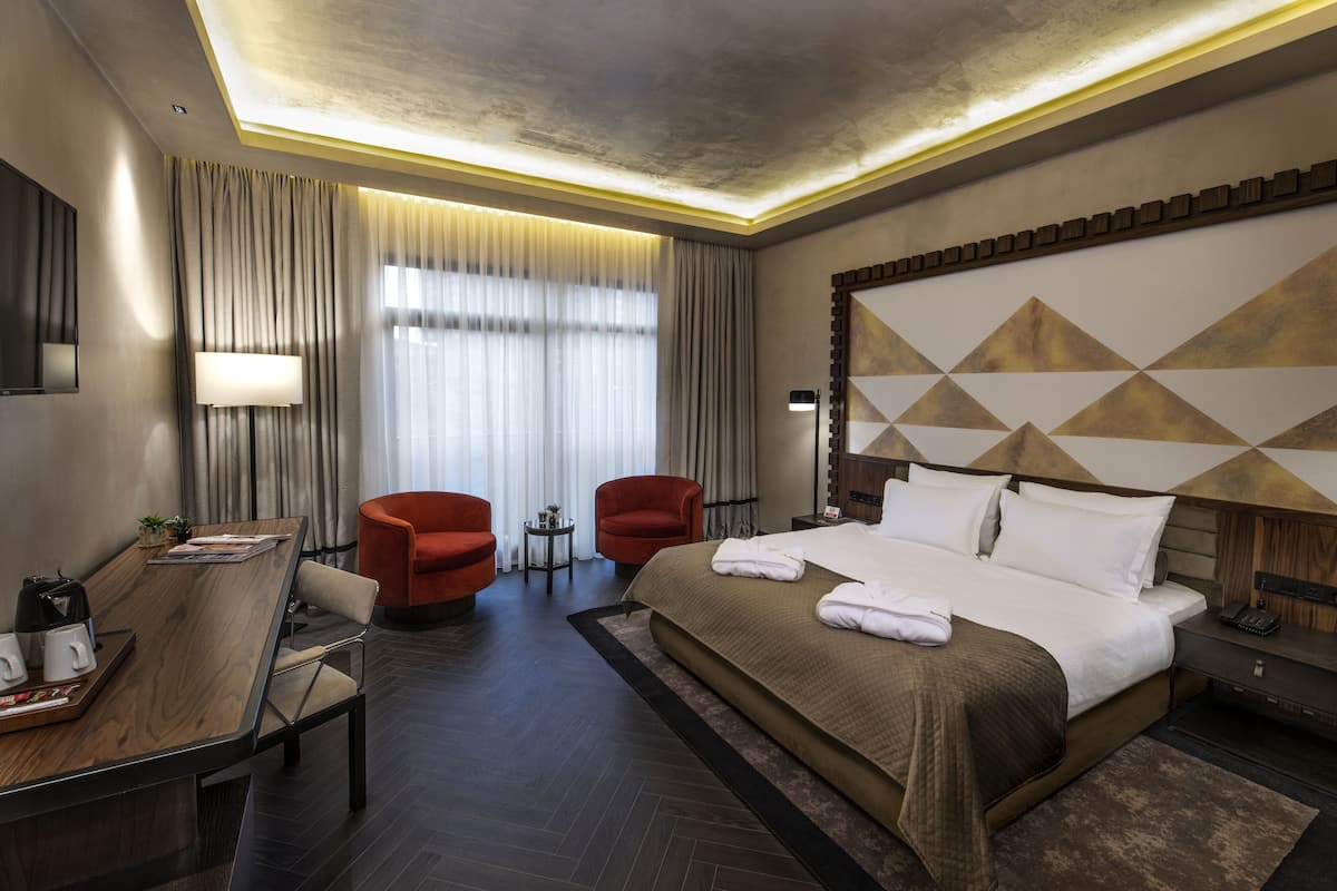 Una habitación de hotel con una cama grande, dos sillones rojos, una mesita de noche con lámpara y un escritorio con cafetera.