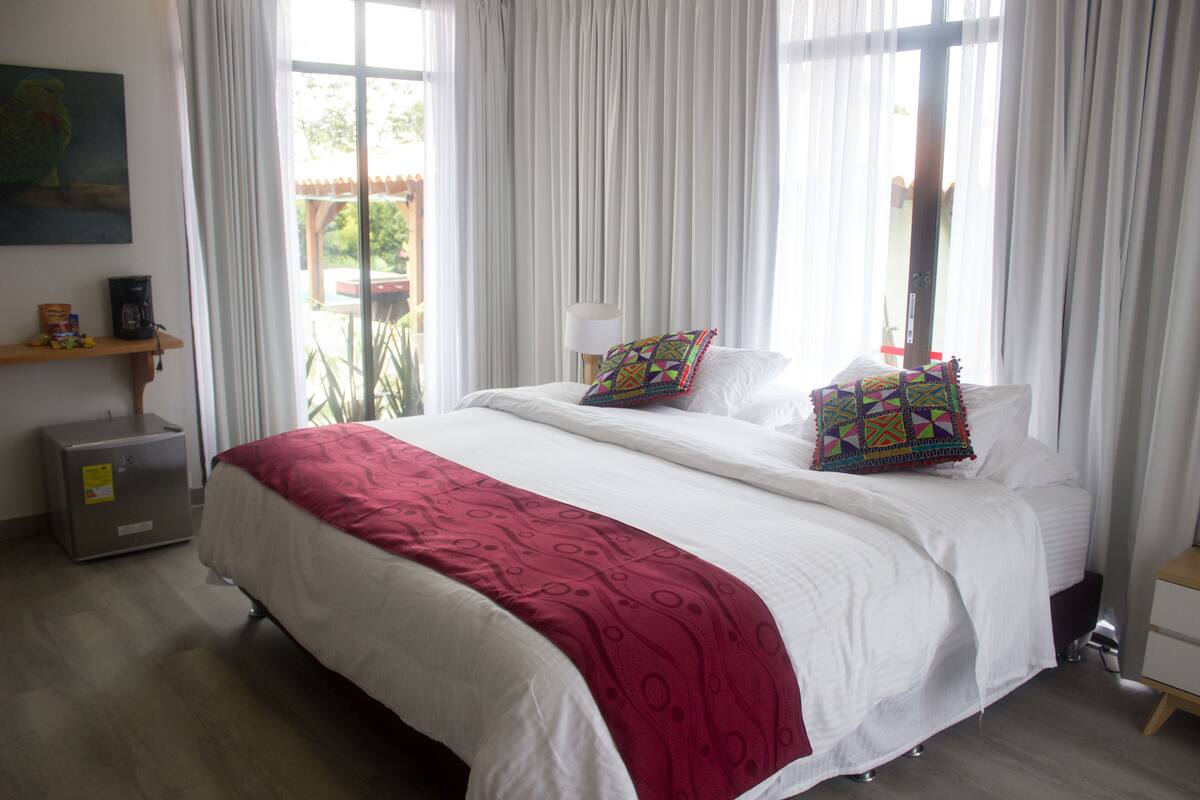 Quarto de hotel com uma cama grande, cortinas brancas, uma pequena mesa de cabeceira, um abajur e vista para o exterior.