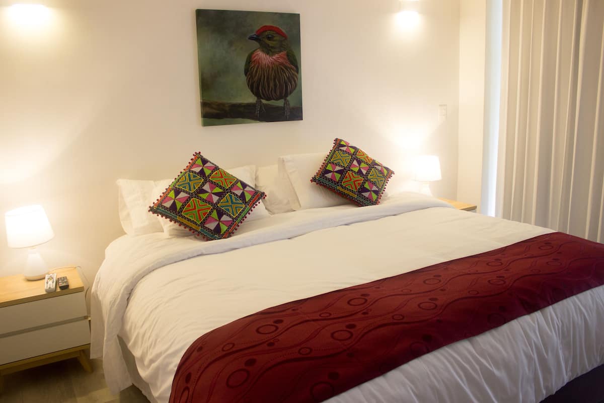 Cama bem arrumada com manta vermelha, duas almofadas coloridas, um quadro de um pássaro na parede e um abajur.