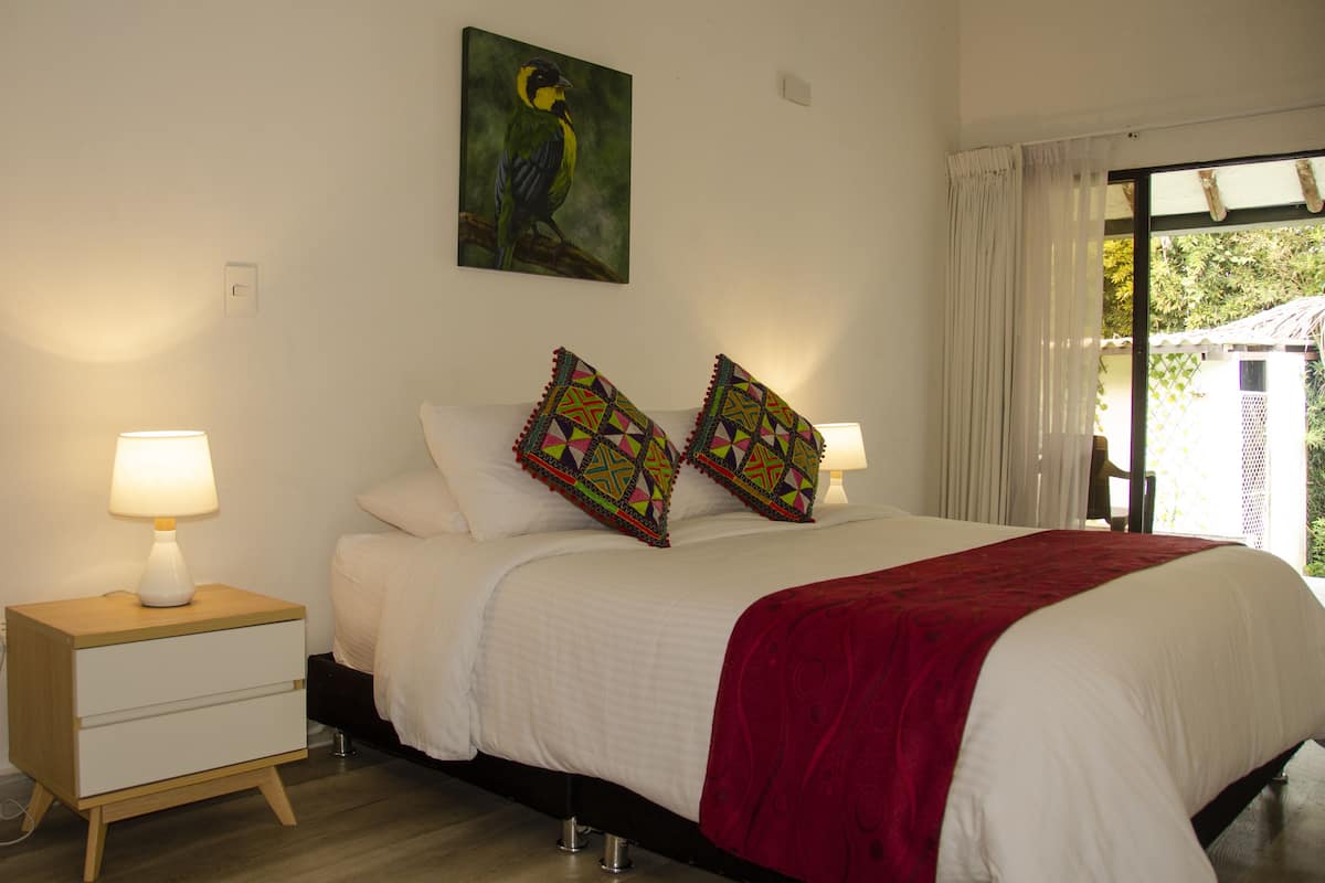 Um quarto com cama, almofadas, mesa de cabeceira, abajur e um quadro na parede.