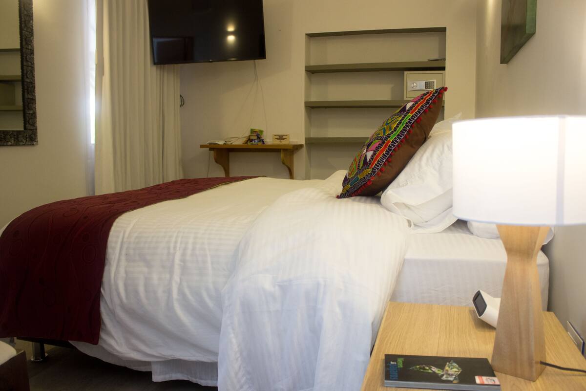 Quarto de hotel com uma cama, uma mesinha de cabeceira com abajur, uma televisão na parede e um criado-mudo de madeira.