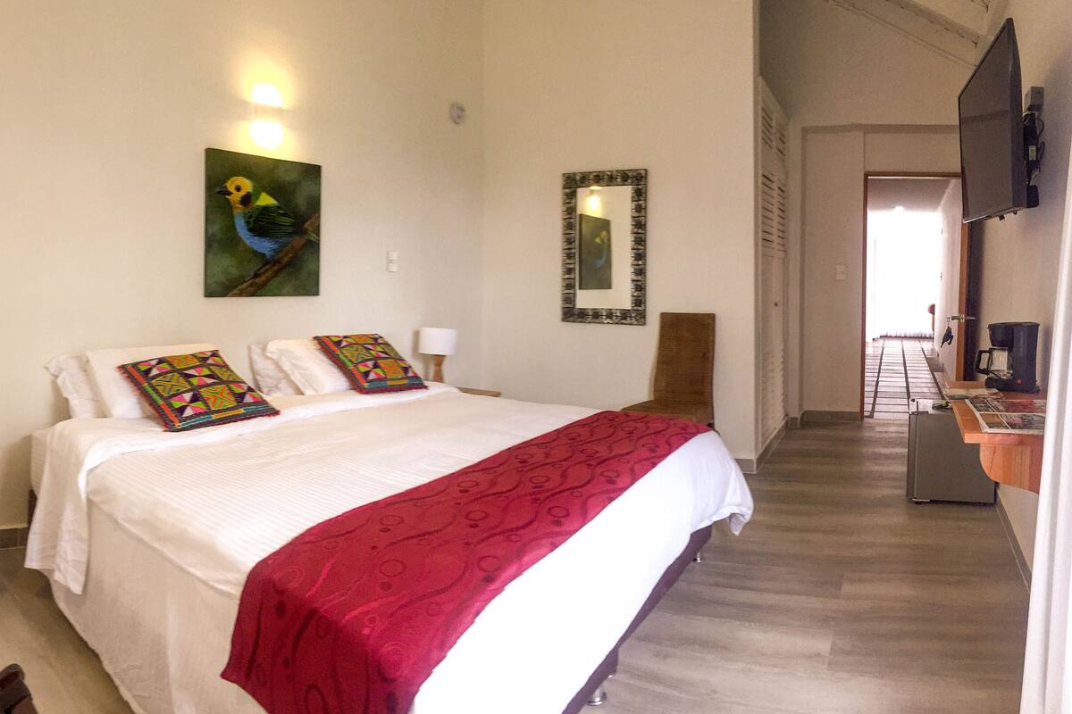 Quarto de hotel com cama, mesa de cabeceira, luminária, quadro, espelho e televisão.