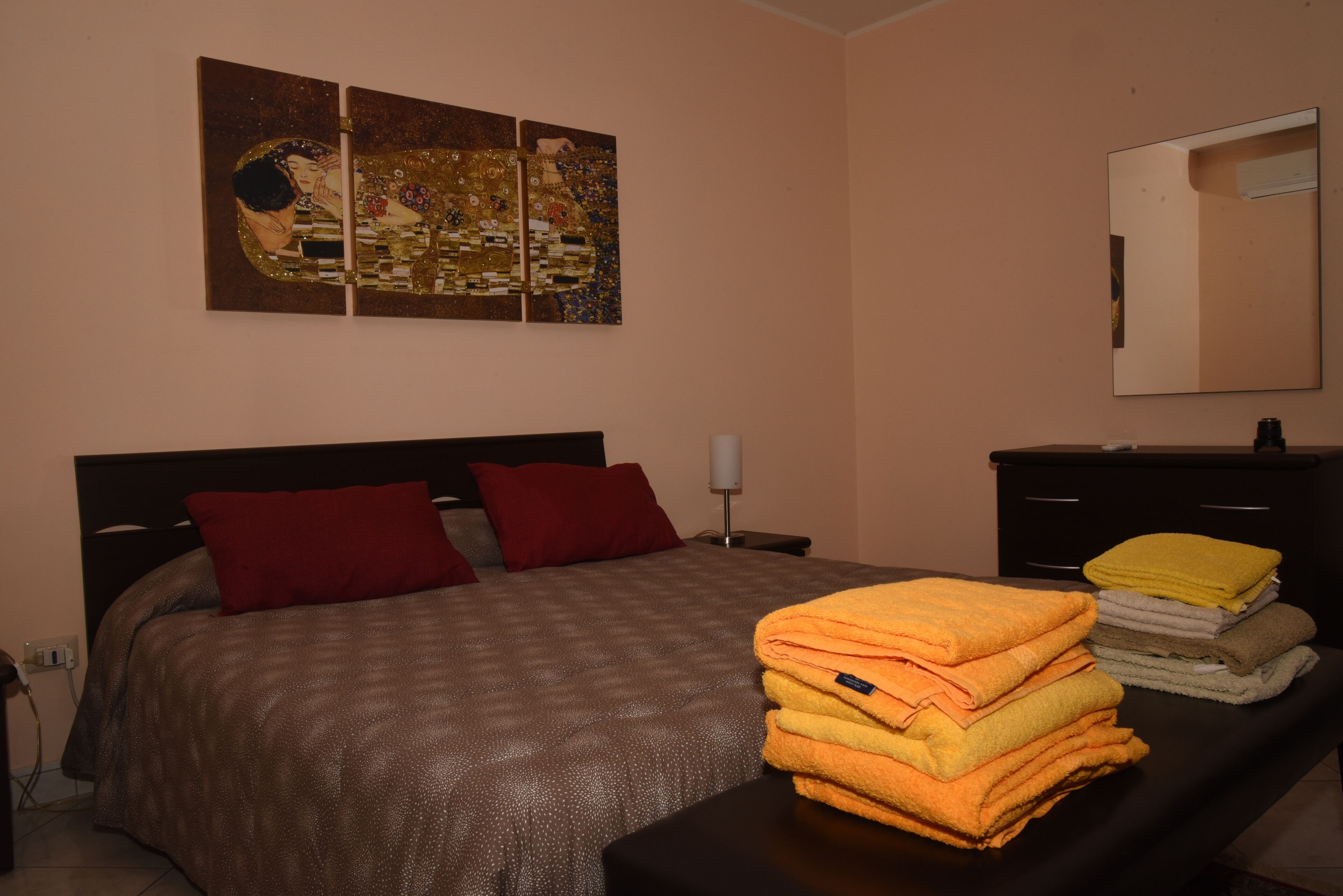 A hotel room with a bed, red pillows, a painting above the bed, a mirror on the wall, and a stack of towels on a table.