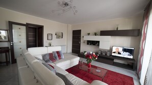 A modern living room with a white sofa, red rug, and a TV displaying a news program. - MODERN AND COZY APARTMENT NEAR THE SEA OF RECANATI (Giardini Naxos)