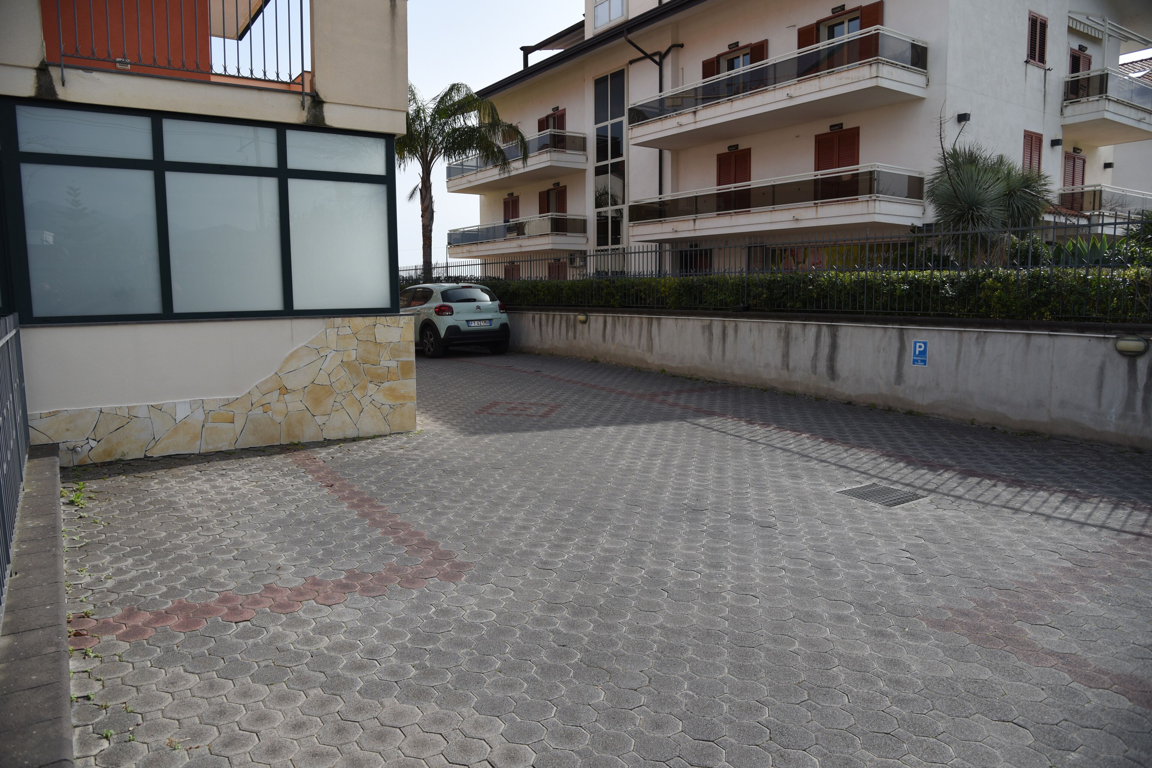 Un'area di parcheggio asfaltata con un'auto parcheggiata, un muro in pietra e un edificio con numerosi balconi e finestre.