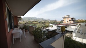 A balcony with a table and chairs, potted plants, and a view of a cityscape and mountains. - MODERN AND COZY APARTMENT NEAR THE SEA OF RECANATI (Giardini Naxos)