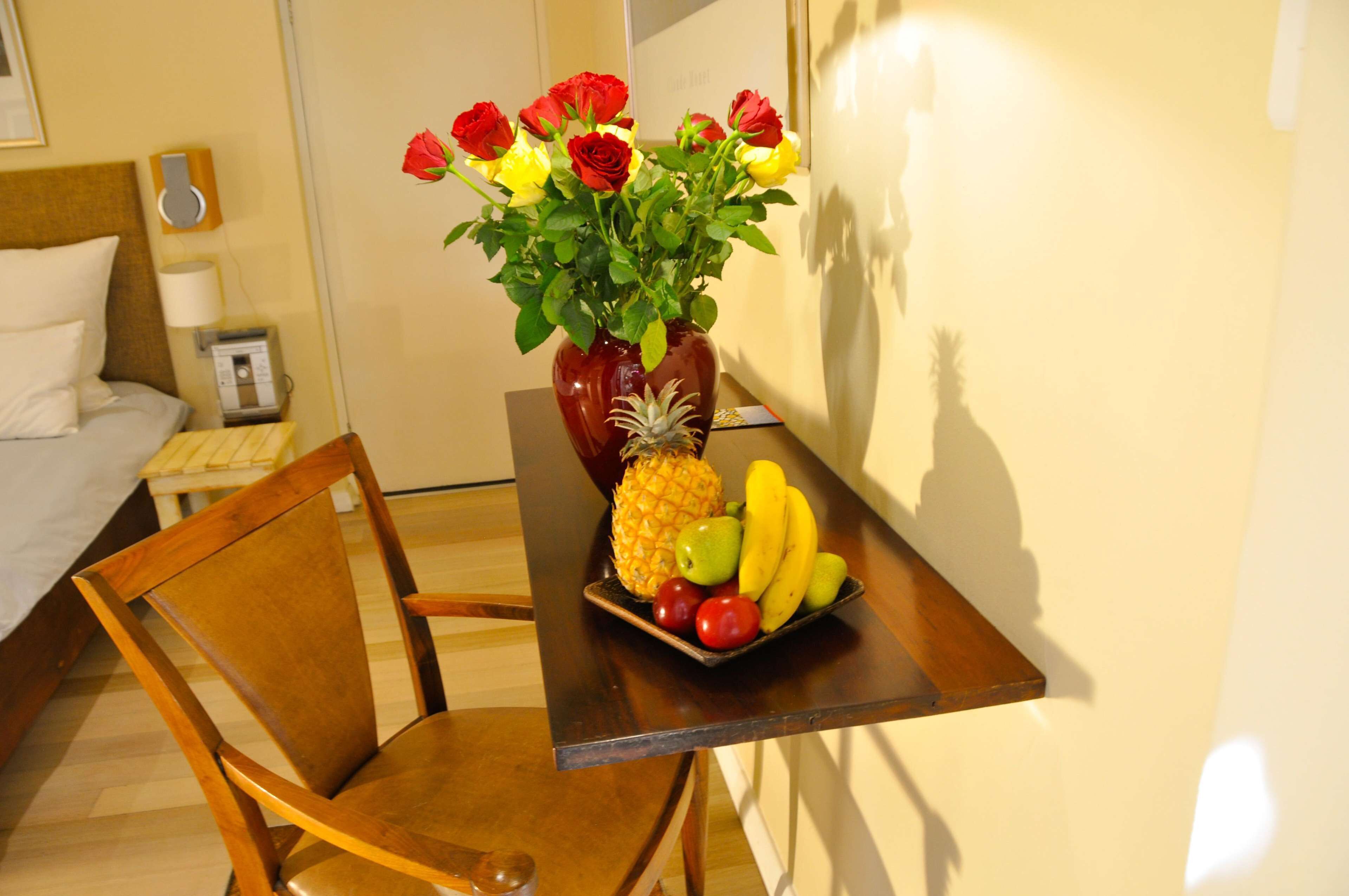 A hotel room with a wooden chair, a small table with a fruit bowl, and a vase with flowers.