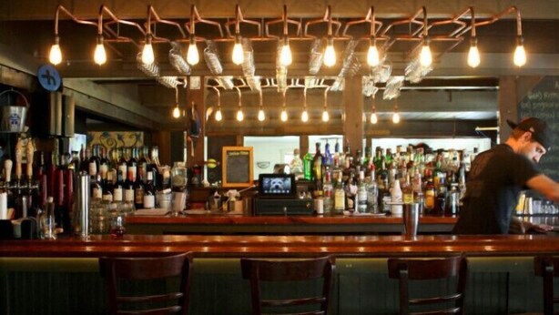 Uma área de bar com cadeiras de madeira, um balcão com diversos tipos de bebidas e um bartender.