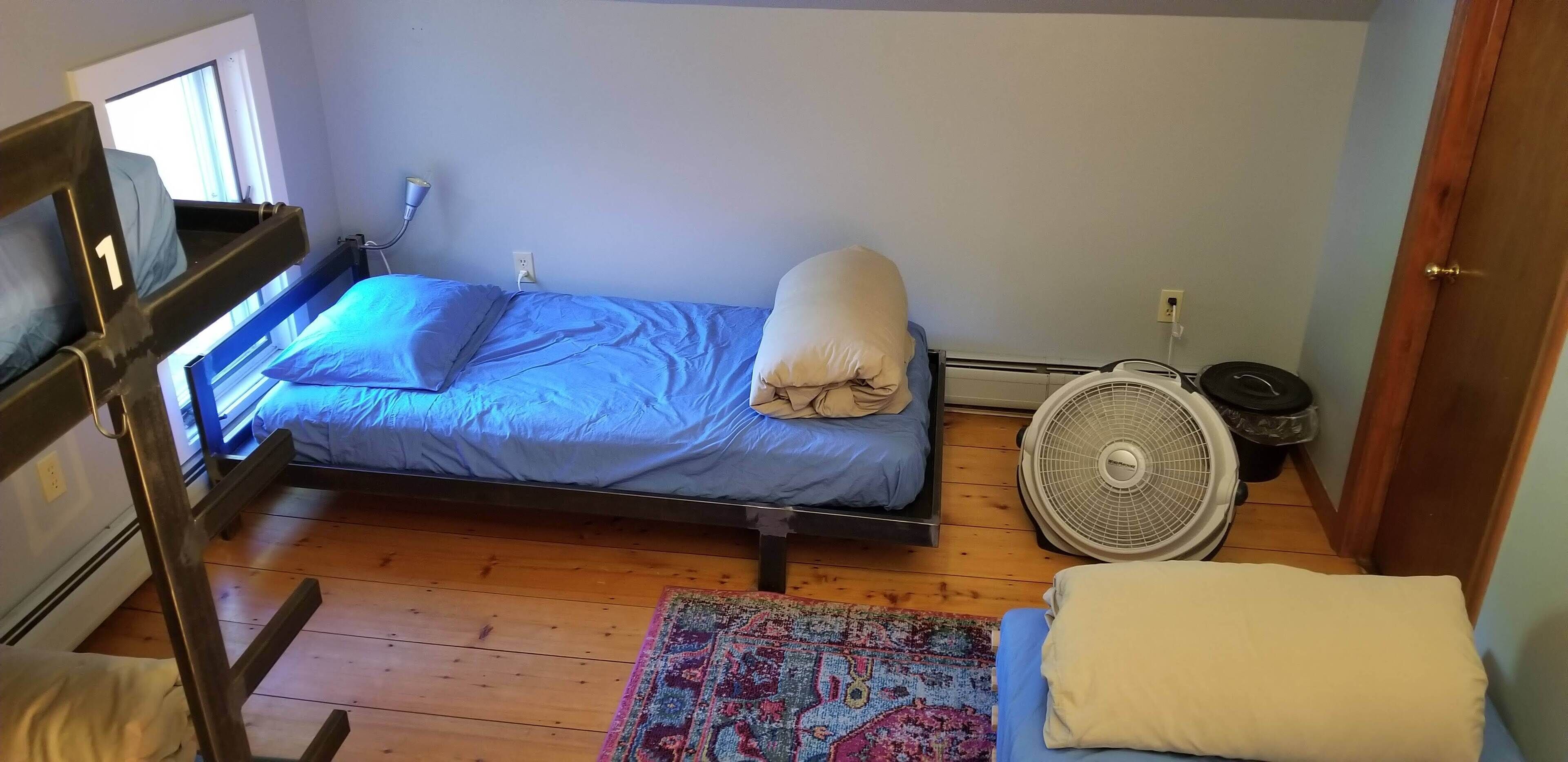 Une petite chambre avec un lit simple, un lit superposé sur le côté gauche, un ventilateur sur pied et une fenêtre avec des rideaux.