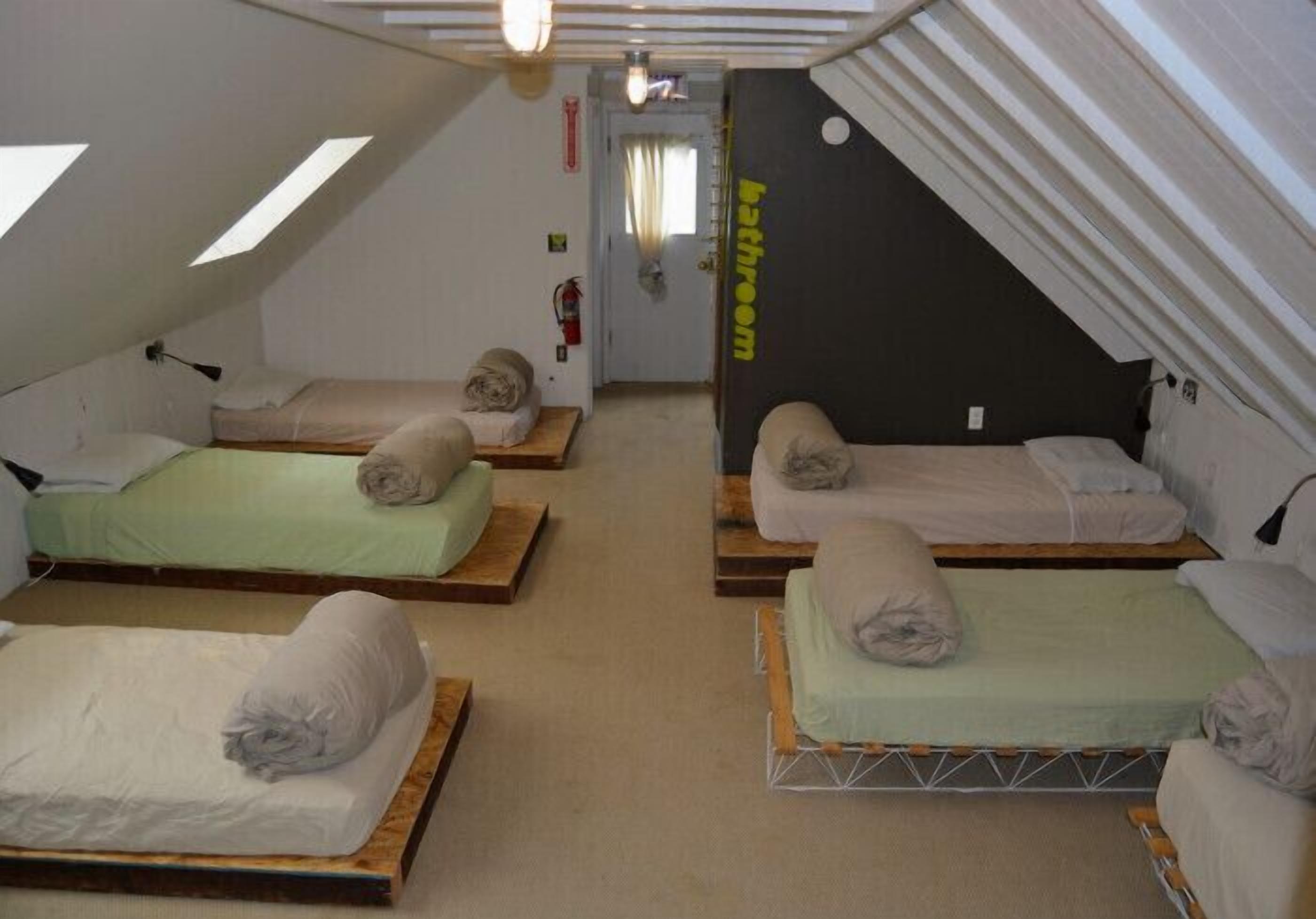 A dormitory-style room with three bunk beds, a visible bathroom door, and a sloped ceiling with skylights.