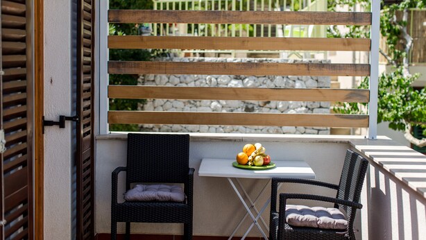 Un balcon agrémenté d’un paravent en lattes de bois, deux fauteuils en osier, une petite table et une assiette de fruits.