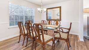 Comedor con mesa de vidrio rodeada de sillas de madera con asientos acolchados, una lámpara de araña, un espejo grande y una ventana con persianas.