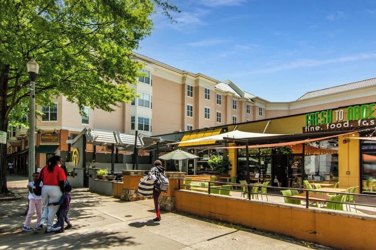 Una calle con mesas al aire libre, un edificio con un letrero que dice 'FRESH TO ORDER' y un árbol que da sombra.