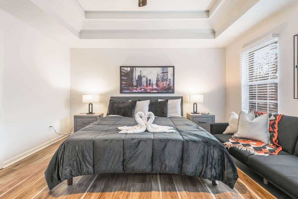 Une chambre moderne avec un grand lit, deux tables de chevet, une œuvre d’art représentant un paysage urbain et un canapé agrémenté d’un plaid à motifs.