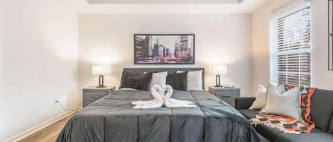A modern bedroom with a large bed, two bedside tables, a cityscape artwork, and a sofa with a patterned throw.