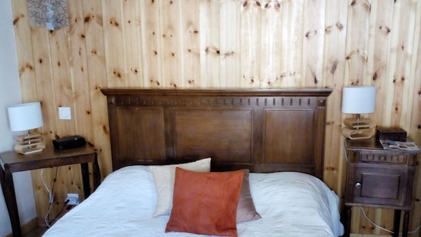 A bedroom with a wooden headboard, a bed with white bedding and a red pillow, two bedside tables with lamps, and a wooden nightstand. - House, large balcony with stairs down to the garden. (L'Argentière-la-Bessée)
