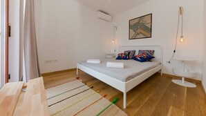 A bedroom with a white bed, colorful pillows, a ceiling fan, an air conditioner, and a painting on the wall.