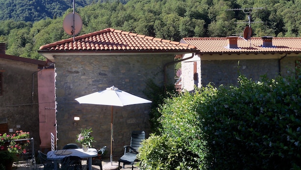 A stone building with a tiled roof, a patio with a table and chairs, and a mountain in the background. - Casa O is a 19th-century, restored, stone house with garden IT046022C2LZ5R4HQX (Pescaglia.)