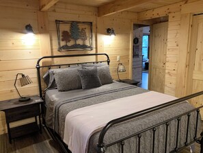 Een slaapkamer in een houten hut met een metalen bed, twee bedlampen, een aan de muur hangend schilderij en een deur die naar een andere kamer leidt.
