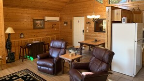 Un accogliente interno di una baita con pareti in legno, un'area pranzo, poltrone in pelle, un frigorifero e una finestra con vista sugli alberi.
