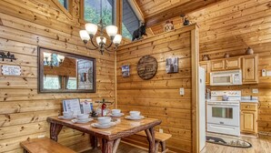 Interno di una baita in legno con un tavolo da pranzo per quattro persone, una cucina con elettrodomestici bianchi e un grande specchio a parete.