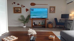 Ein Wohnzimmer mit Fernseher, der eine Strandansicht zeigt, einem Holztisch, einer Fernbedienung, einer Topfpflanze und einem gerahmten Bild an der Wand.