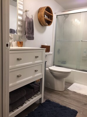Un baño con inodoro, lavamanos con espejo, estante de madera y alfombra azul oscuro.