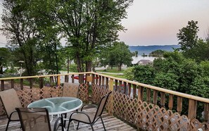 A wooden deck with a glass table and chairs, overlooking a lake and a road.