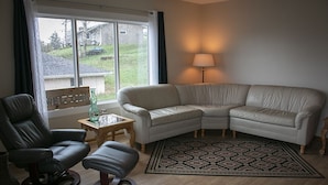 Ein Wohnzimmer mit einem Ecksofa, einem schwarzen Ledersessel, einem kleinen Tisch mit einer Flasche und einer Lampe.