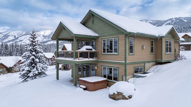 A multi-story lodge with a balcony, a hot tub, and a view of snow-covered mountains.
