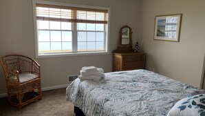 A bedroom with a large window, a wooden dresser, a wicker chair, and a bed with a floral bedspread.