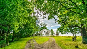 A white house with a grey roof, surrounded by lush green trees and a dirt path leading to it.