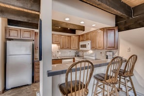 A kitchen with wooden cabinets, a refrigerator, a microwave, and a stove. There are three bar stools and a sink.