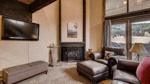 A living room with a fireplace, leather sofas, a flat-screen TV, and a large window with a view of a forest. - Perfect Ski In/Ski Out Condo in Center Village (Copper Mountain)