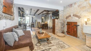 A cozy living room with a brown sofa, a wooden coffee table, and a stone wall. There is a kitchen area with modern appliances and a dining table with chairs.