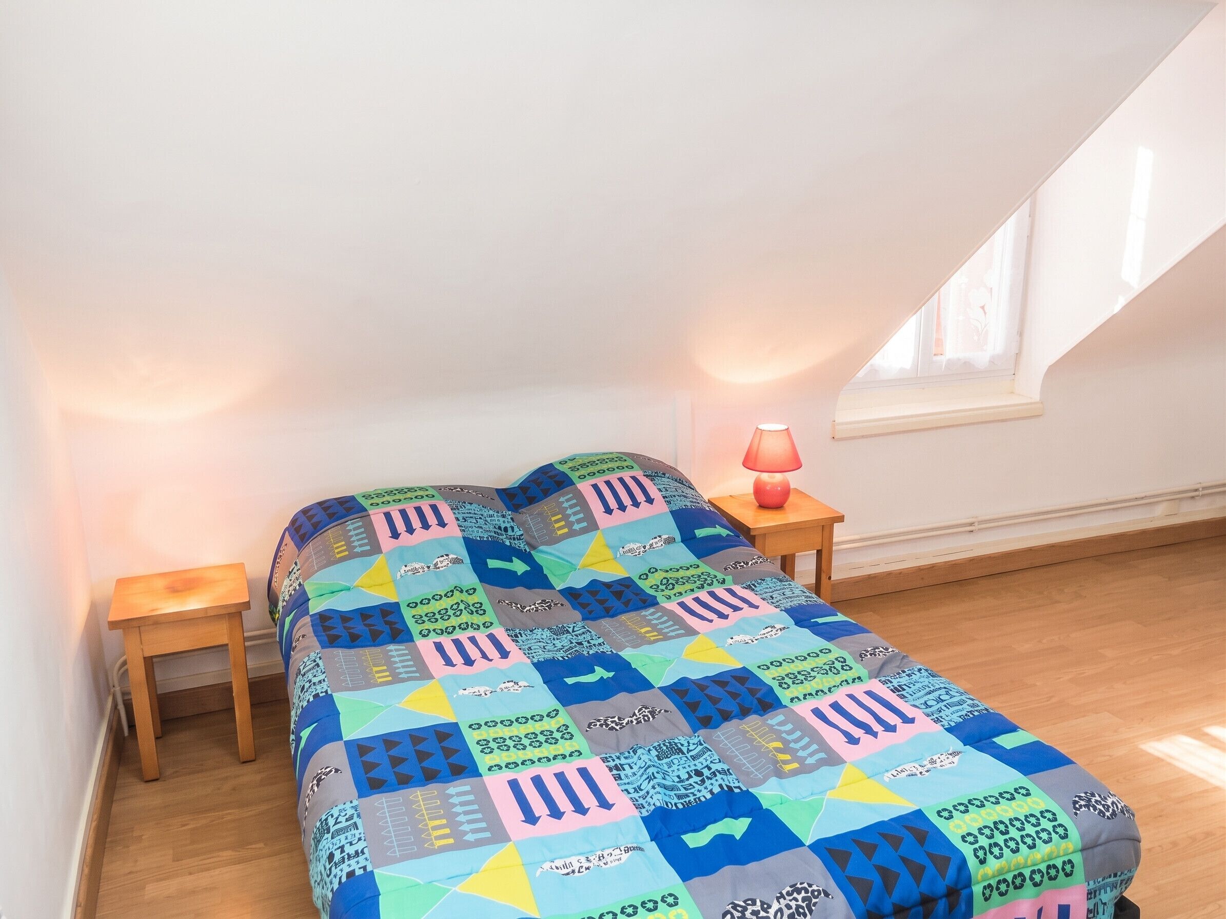 A bedroom with a colorful quilt, a bedside lamp, and a window.
