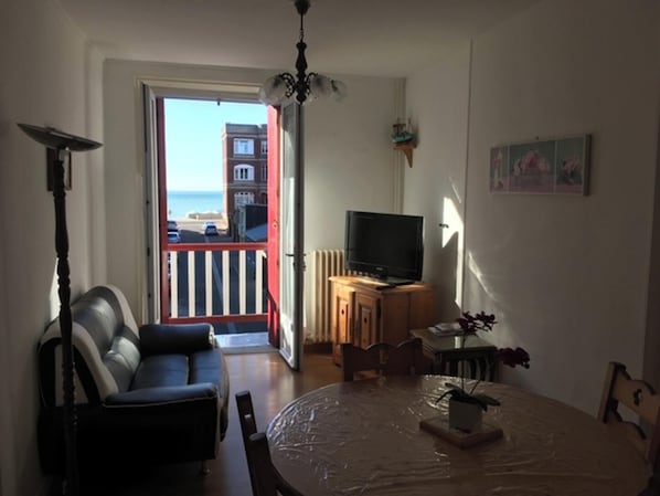 Un salon avec un canapé, un téléviseur posé sur un meuble en bois, une table à manger recouverte d’une nappe en plastique et une vue sur la mer par une porte-fenêtre donnant sur le balcon.